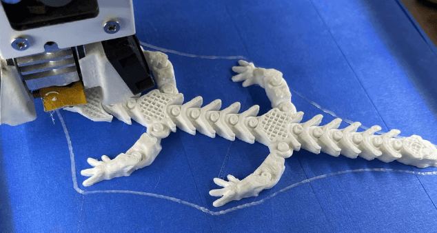 3D printer producing a student-designed white articulated lizard model on a blue print bed