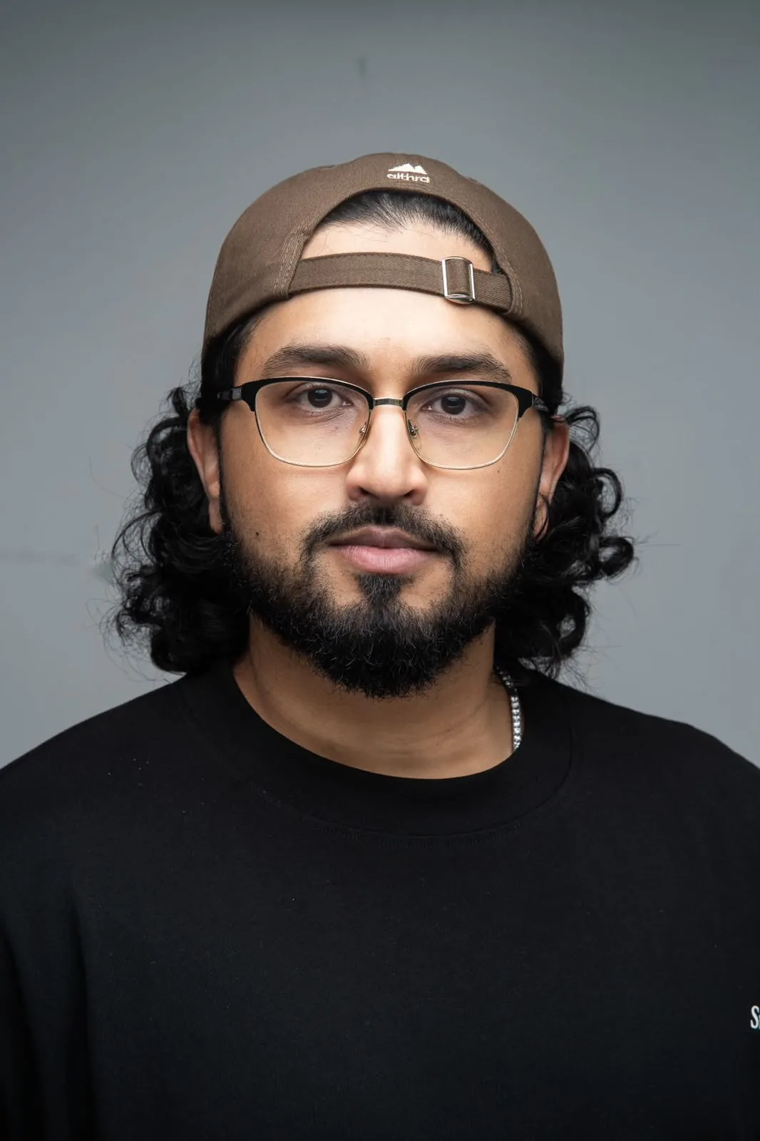 Kirat P, Business and Digital Media Instructor, in a backwards brown cap and glasses against a grey wall