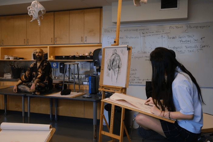 Life-drawing class with a costumed model posing beside a student's easel study
