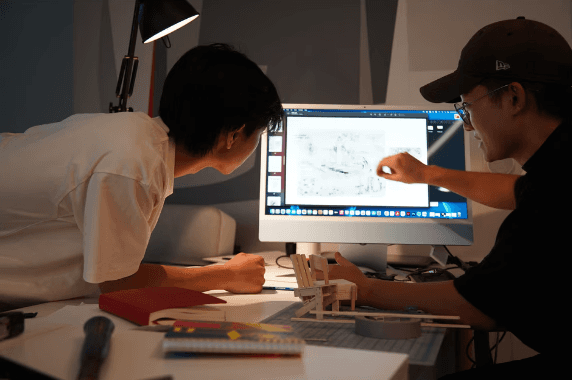 Mentor and student reviewing drawings together on a desktop monitor beside a wooden model