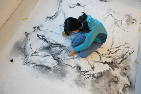 Student kneeling on a room-sized charcoal drawing, working directly on the paper