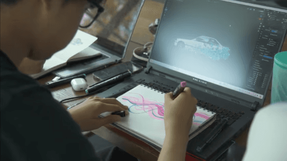 Student drawing concept sketches in a notebook beside a laptop showing a 3D car model
