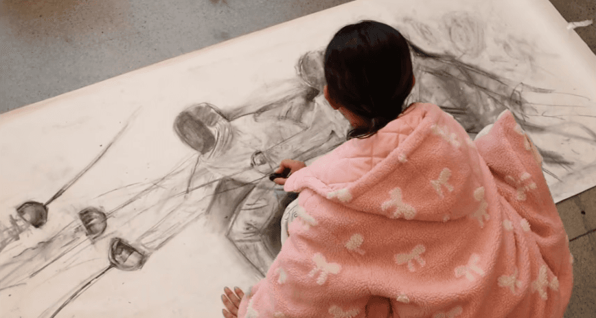Young student in a pink hoodie working on a large charcoal figure drawing spread across the floor