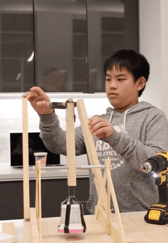 Young student assembling a wooden kinetic structure on a worktable with a power drill