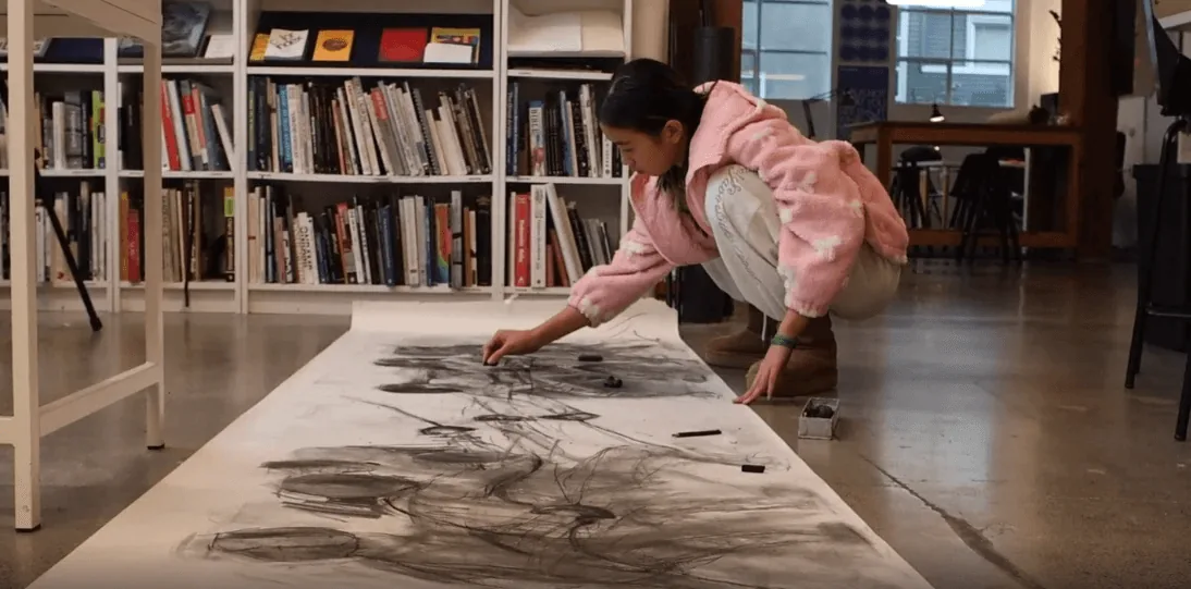学生越过长长的工作室桌面，在书架旁创作一幅大型炭笔画