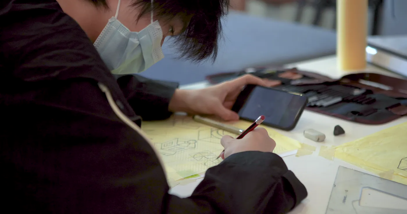 Student sketching designs on paper from a reference photo on their phone