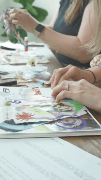 Students collaging cut-paper compositions into sketchbooks at a shared worktable