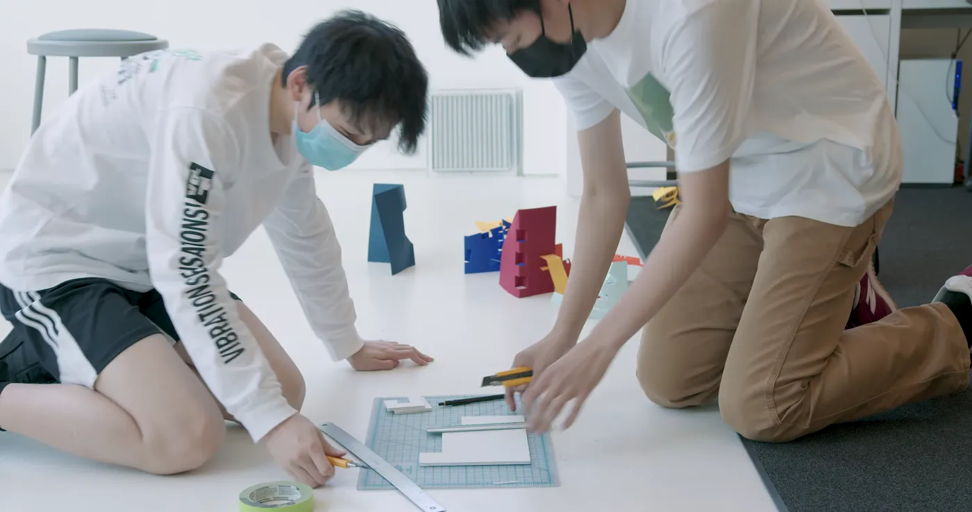 Two students measuring and cutting parts for a scale model on the studio floor
