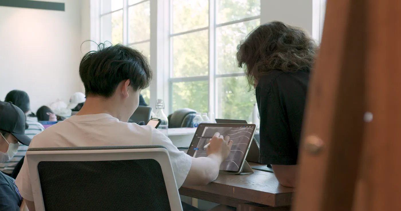 Two students drawing on tablets at a worktable beside the studio windows